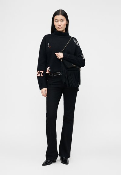 Femme debout portant un pull à col roulé noir avec des patchs, un pantalon évasé noir, des chaussures noires pointues et tenant un sac à bandoulière noir à franges.