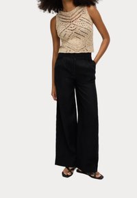 Gehaakte tanktop in beige met ingewikkelde patronen, gecombineerd met zwart hoge tailles brede broek. Zwarte platte sandalen maken de outfit compleet.