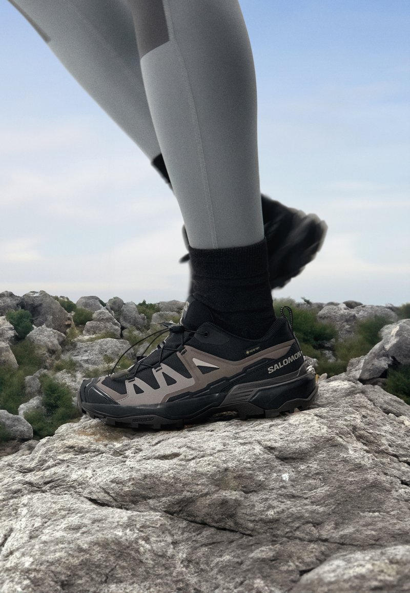 Svart og grå utendørssko med teksturert såle, perforert overdel og snøresystem, brukt på en steinete overflate, komplett med grå leggings.