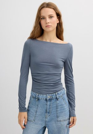 Top de manga larga en tejido azul suave con escote barco y detalles fruncidos en los laterales, combinado con pantalones vaqueros claros de cintura alta.