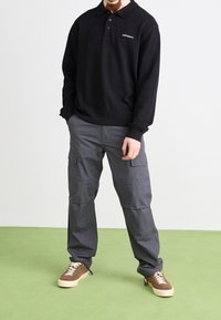 Man in zwart lange mouwen Carhartt polo, grijze cargo broek en bruine sneakers, staande op groene vloer tegen een witte muur.