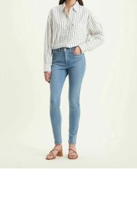 Vaqueros ajustados de cintura alta de un azul claro en denim. Combinados con una camisa de rayas de manga larga blanca y sandalias de tacón bloque en color beige.