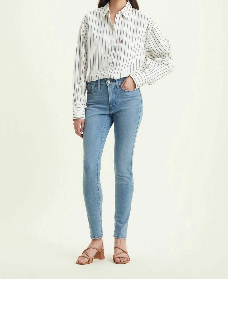 Vaqueros ajustados de cintura alta de un azul claro en denim. Combinados con una camisa de rayas de manga larga blanca y sandalias de tacón bloque en color beige.