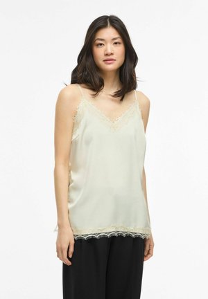 Kobieta z ciemnymi włosami ubrana w kremowy top z koronką i czarne szerokie spodnie na białym tle.