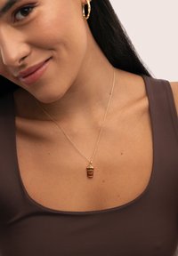 Collar tono dorado con un colgante de taza de café, que presenta grabado "DÍA DEL CAFÉ", sobre una delicada cadena, con un fondo marrón.