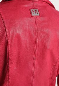 Czerwona skórzana kurtka o gładkim wykończeniu, z teksturowanymi siateczkowymi detalami po bokach oraz kwadratową naszywką z logo przy kołnierzu.