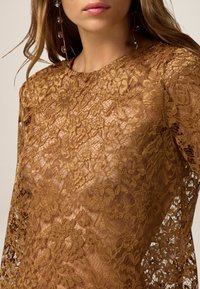 Top in pizzo di un ricco colore marrone, caratterizzato da motivi floreali, maniche lunghe e una finitura semi-trasparente, con dettagli intricati e una texture raffinata.