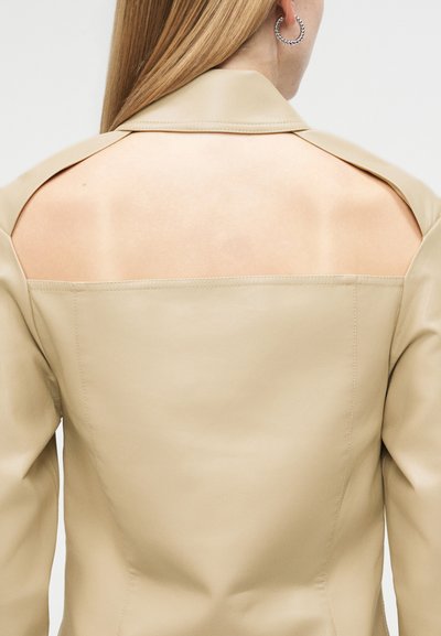 Žena s rovnými blond vlasmi nosí béžovú koženú bundu s výrezom na chrbte, ktorý odhaľuje hornú časť chrbta a plecia, a striebornú kruhovú náušnicu.