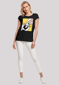 Svart t-skjorte med korte ermer som har et fargerikt grafisk trykk av en tegneseriekanin på gul bakgrunn. Kombinert med hvite skinny jeans og hvite sneakers.