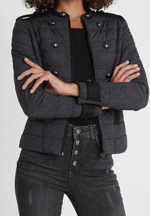 Woman wearing a dark gray quilted military-style jacket with black buttons, black shirt, and high-waisted dark jeans with four buttons.