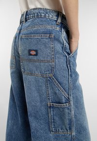 Eine Nahaufnahme von Jeanshosen mit einer großen Tasche, die ein Dickies-Logo auf der Rückseite zeigt. Die Person hat eine Hand in ihrer Tasche.
