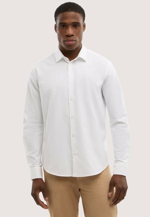 Mężczyzna ubranay w białą koszulę z długim rękawem zapinaną na guziki oraz beżowe spodnie, stojący na tle jednolitego jasnego tła.