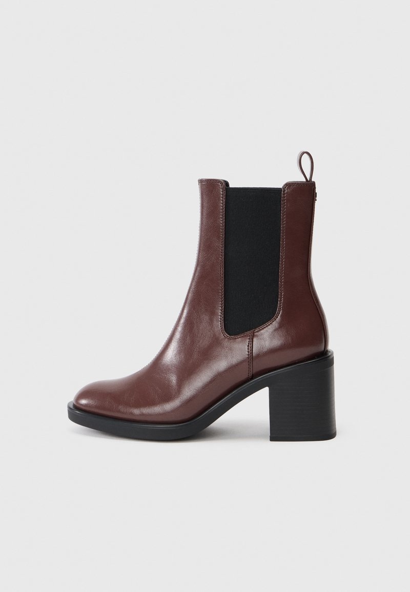 Bottine en cuir marron avec panneau latéral élastique noir, talon bloc, bout arrondi et languette arrière sur fond gris clair.