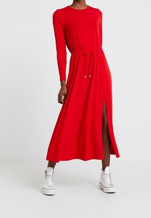 Femme portant une longue robe rouge avec une taille à cordon et une fente latérale, associée à des baskets blanches montantes, sur un fond uni.