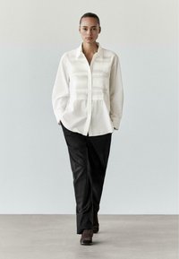 Bílá košile s knoflíky a texturovanými sklady vpředu, v kombinaci s černými kalhotami a hnědými kotníkovými botami, na neutrálním pozadí.