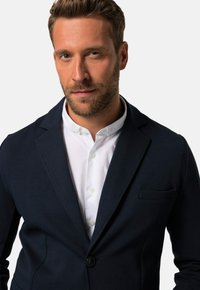 Navy Blazer aus strukturiertem Stoff mit einem Ein-Knopf-Verschluss und einer Brusttasche, getragen über einem weißen Hemd.
