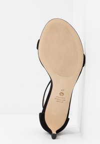 Chaussure à talon haut en suède noir, inclinée pour montrer la semelle beige avec une zone de grip texturée et le texte de la marque "Stuart Weitzman, Fabriqué en Espagne, taille 37."