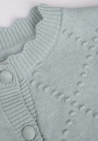 Jasnoniebieski sweter z dzianiny z prążkowanym kołnierzem, dekoracyjną fakturą i okrągłymi guzikami. Posiada falbaniasty brzeg oraz małe wypukłe wzory.