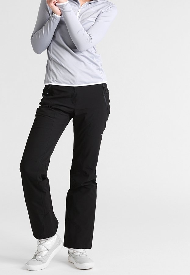 Personne portant un haut gris clair à manches longues, un pantalon noir et des chaussures gris clair, debout les bras croisés sous le visage sur un fond gris.