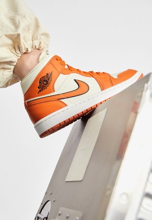 Pomarańczowo-kremowe wysokie sneakers Air Jordan noszone z beżowymi spodniami, spoczywające na srebrnym metalowym pudełku na białym tle.