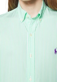 Henkilö, jolla on yllään napitettava vihreä ja valkoinen pystyraitapaita, jossa on napitettavat kaulukset ja pieni violetti polo-pelaajan logo rinnassa.