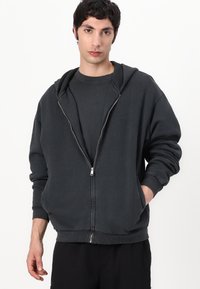 Black zip-up hoodie made of cotton, featuring a hood, ribbed cuffs, and front pockets. Paired with a matching dark shirt underneath.