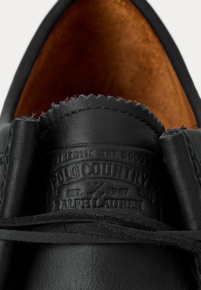 Prim-plan al limbii unui pantof negru din piele, cu sigla Polo Country Ralph Lauren embosată și interiorul pantofului din piele maro vizibil.
