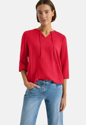 Femme portant un chemisier rouge vif à col en V avec cordons et manches 3/4, associé à un jean taille haute bleu clair avec double bouton.
