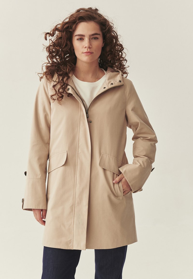 Žena s kučeravými vlasmi, oblečená v béžovom dlhom kabáte cez bielu košeľu a tmavé nohavice, so z rukou vrecku kabáta, stojaca pred jednofarebným pozadím.