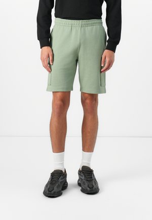 Shorts en coton vert clair avec une taille élastique, des poches latérales et une coupe décontractée, associés à des baskets noires et des chaussettes blanches.