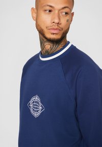 Mørkeblå sweatshirt med hvid ribbet krave, der har et hvidt geometrisk broderet design på brystet. Blødt, tekstureret stof.