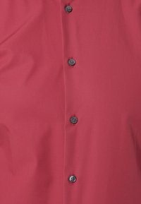 Rotes Hemd mit glatter Textur und schwarzen Knöpfen entlang der Vorderseite. Der Stoff zeigt leichte Falten, was auf Weichheit hinweist.