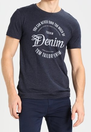 Mężczyzna ubrany w ciemnoniebieski t-shirt z białym napisem o denym i granatowe spodnie, stoi z rękami swobodnie opuszczonymi wzdłuż ciała.