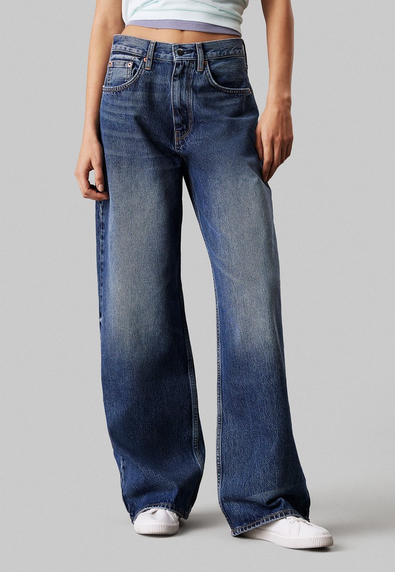 Wijduitlopende denim jeans in donkerblauw met lichte vervaging, vijf-pocket ontwerp en een sluiting met knoop aan de voorkant. Gecombineerd met witte sneakers.