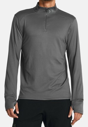 Mężczyzna noszący szary sportowy top z długim rękawem, zamkiem do ćwiartki i otworami na kciuki, w połączeniu z czarnymi spodniami sportowymi.