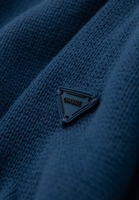 Blau gestrickter Stoff mit einem strukturierten Muster, das ein dreieckiges schwarzes Etikett mit dem eingeprägten Wort "OMBRE" zeigt.