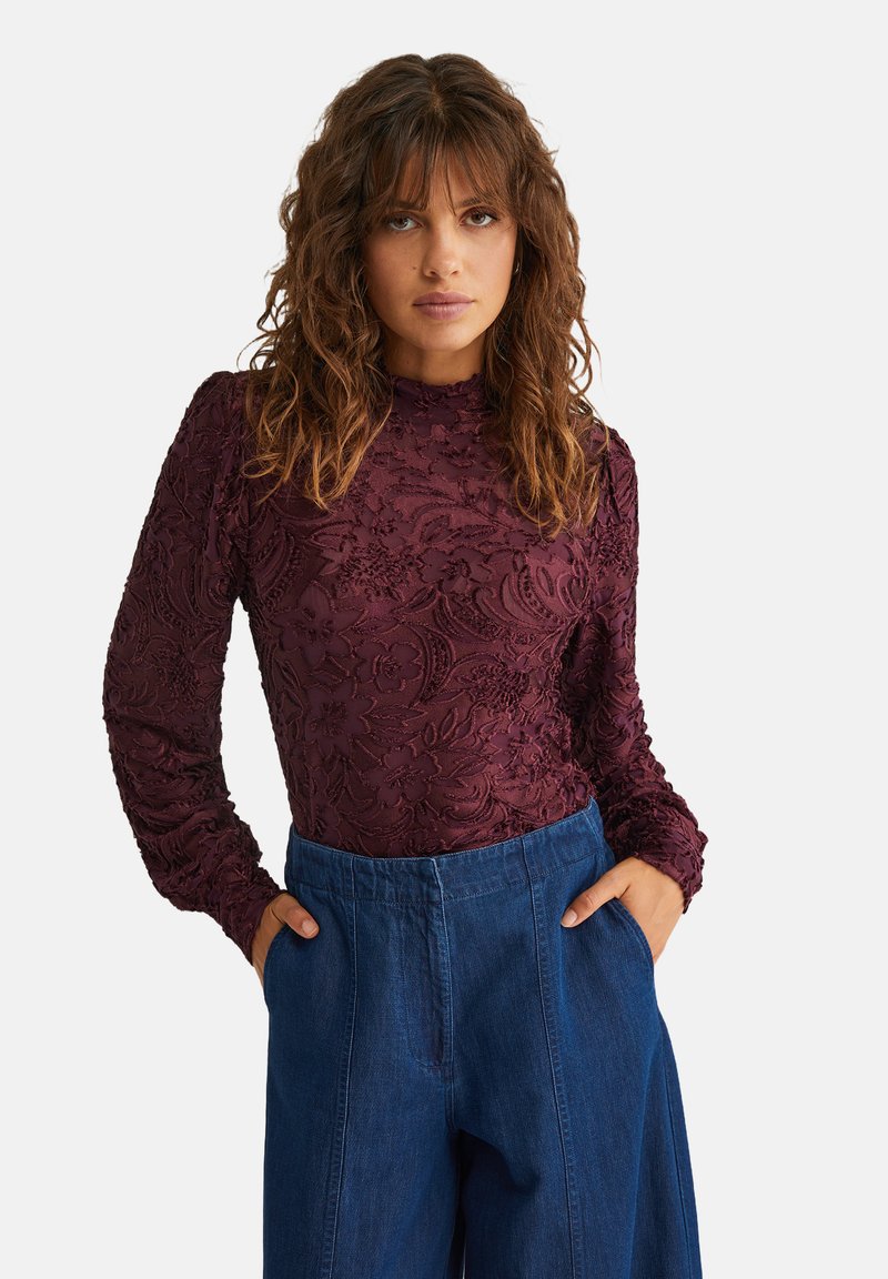 Top lung cu mâneci lungi de culoare rosu închis, cu un design floral texturat și guler înalt, asortat cu pantaloni din denim cu talie înaltă.