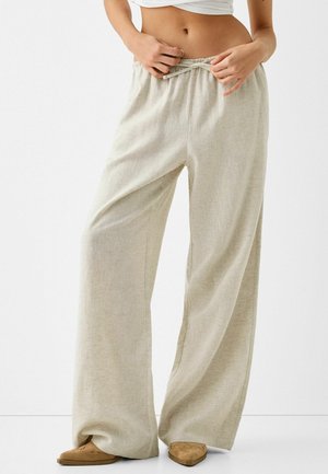 Personne ajustant le cordon de pantalons amples en lin beige, portant des chaussures marron pointues et un top court blanc, debout devant un fond uni.