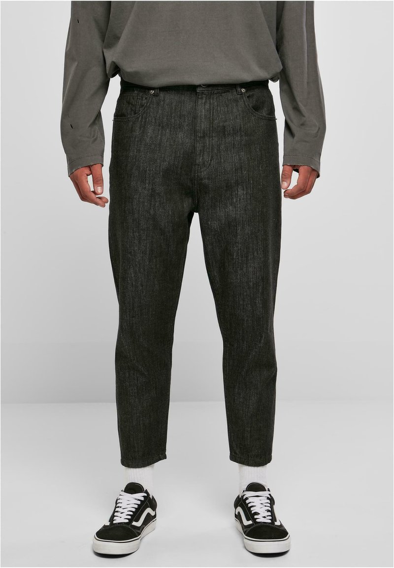 Schwarze Jeans aus Denim mit geradem Schnitt, ausgestattet mit fünf Taschen, einem Reißverschluss und subtiler Textur. Getragen mit schwarzen Sneakers und weißen Socken.