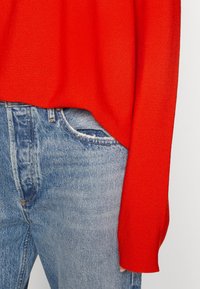 Roter Strickpullover mit langen Ärmeln, kombiniert mit hellblauen, ausgewaschenen Jeans, die eine Fronttasche und eine strukturierte Stoffoberfläche aufweisen.