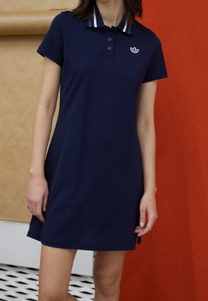 Femme portant une robe polo à manches courtes bleu marine avec un col à rayures blanches et un logo Adidas sur la poitrine, debout devant un fond rouge et beige.