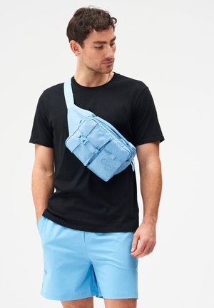 Mann trägt schwarzes T-Shirt und hellblaue Shorts mit passender hellblauer Umhängetasche, schaut nach unten links.