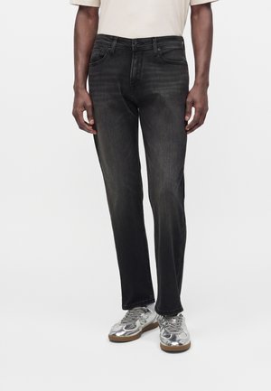 Mand iført falmede sorte straight-leg jeans og metallic sølv sneakers står mod en ensfarvet hvid baggrund.