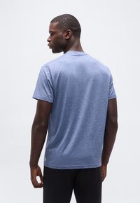 Άνδρας που κο ιτάζει προς τα πίσω, φοράει μπλε ριγέ κοντομάνικο μπλουζάκι και μαύρο παντελόνι, με απλό ανοιχτό φόντο.