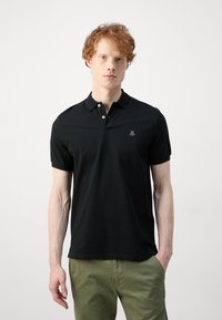 Camisa polo negra de manga corta, con tapeta de dos botones y logo bordado en el lado izquierdo del pecho. La tela parece suave y elástica.