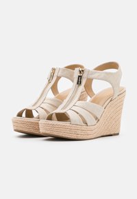 Sandalias de cuña en tejido beige claro con los dedos al aire. Cuentan con una cremallera frontal y plataformas de yute trenzado, creando un contraste texturizado.