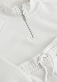 Sweat-shirt blanc à fermeture éclair avec texture côtelée, doté d'un col montant, d'une fermeture éclair argentée et d'un cordon de serrage ajustable à l'ourlet.