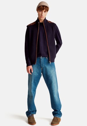 Gilet a maglia blu navy scuro con rifiniture marroni sulle spalle, maglione blu navy sotto, jeans blu e scarpe in suede marroni, indossato con un berretto a maglia beige.