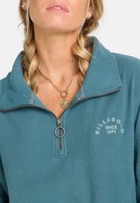 Tyrkysový zip-up pulovr vyrobený z žebrované látky, s límečkem a vyšitým logem "Billabong". Viditelný řetízek s přívěskem.