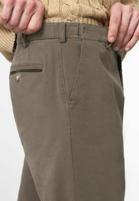 Chemise en tissu beige, courte, montrant un pantalon vert-brun avec un devant plat, une poche latérale et un accent bouton sur la poche arrière. Texture lisse.
